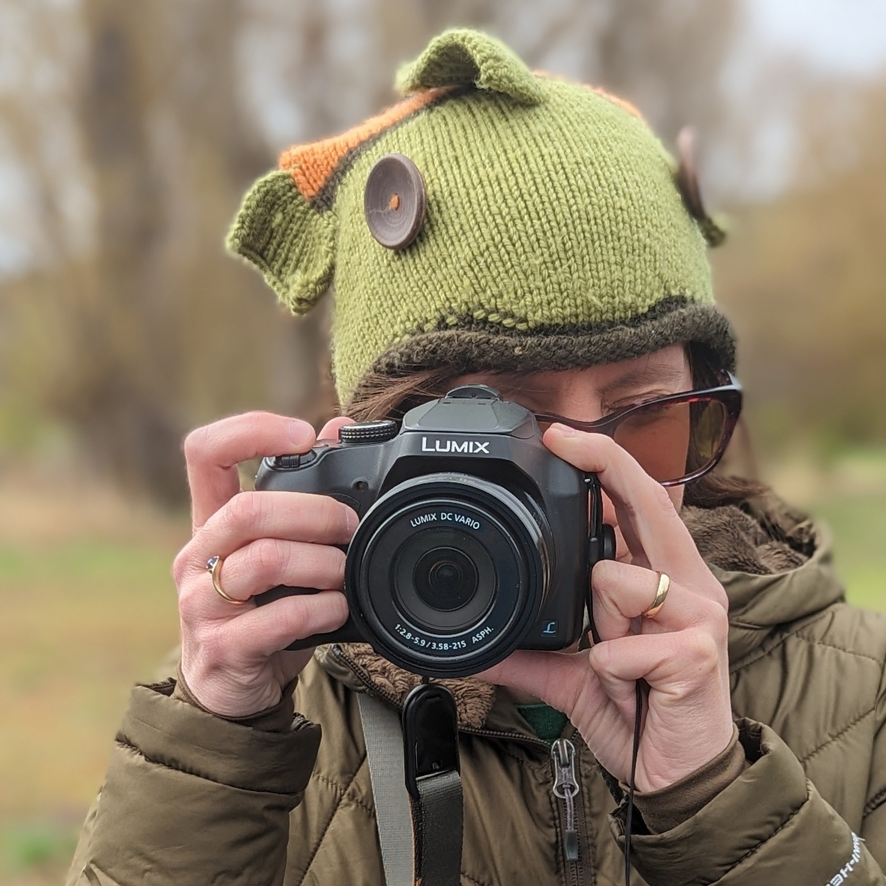 close-up profile pic of a light-skinned person with messy brown hair wearing a colorful knit cap shaped like a fish and holding up a camera as though taking your picture