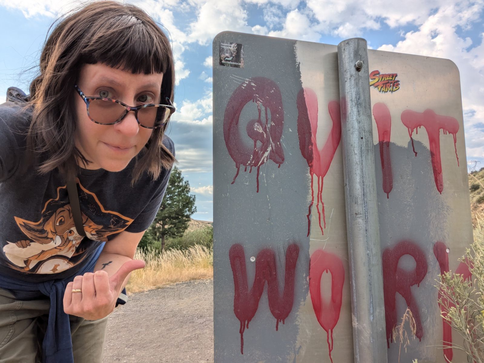 photo of a light-skinned person with wavy, shoulder-length brown hair and colorful tinted glasses and a raven tattoo; she/they are leaning over to gesture emphatically at a trailhead sign that has been tagged with graffitti that reads Quit Work